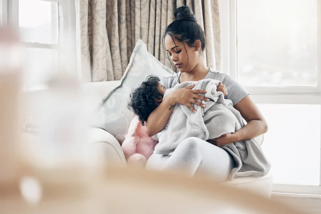 Mother sitting by a window holding her young child, who is wrapped in a gray blanket.